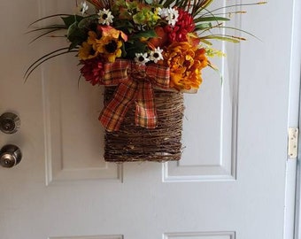 May include: A decorative floral arrangement in a woven basket, hanging on a white door. The arrangement features orange, yellow, and red flowers, with green foliage and a plaid ribbon. The door has a decorative arched window.