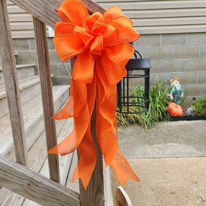 May include: A large orange bow tied to a wooden railing. The bow is made of a sheer fabric and has a large, fluffy center. The bow is tied to a wooden railing, which is part of a set of stairs.