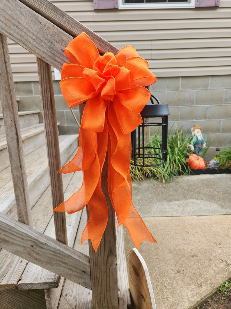 May include: A large orange bow tied to a wooden railing. The bow is made of a sheer fabric and has a large, fluffy center. The bow is tied to a wooden railing, which is part of a set of stairs.