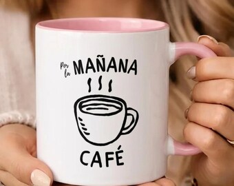 May include: White ceramic coffee mug with a pink interior and handle. The mug features the words "Por la Mañana Café" in black, along with a black illustration of a coffee cup with steam rising from it. The mug is held by a person.