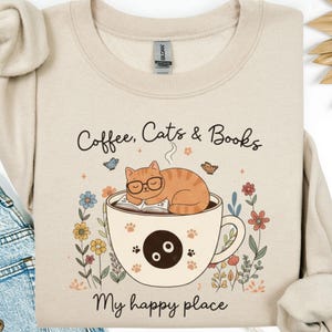 Puede incluir: Sudadera beige con el texto "Coffee, Cats & Books" y "My happy place". El diseño presenta un gato con gafas durmiendo en una taza de café con un libro. Acentos florales y mariposas rodean la taza.