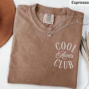 Puede incluir: Camiseta color espresso con el texto "COOL AUNTS CLUB" en escritura blanca. La camiseta tiene cuello redondo y mangas cortas. Un collar plateado con un pequeño colgante está drapeado sobre la camiseta.