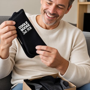 May include: A pair of black crew socks featuring the white printed text COOLE SOCKE Seit 1976. The socks are made of a ribbed fabric and are presented as a gift, accompanied by a small card with the handwritten text Happy Birthday and a heart symbol.