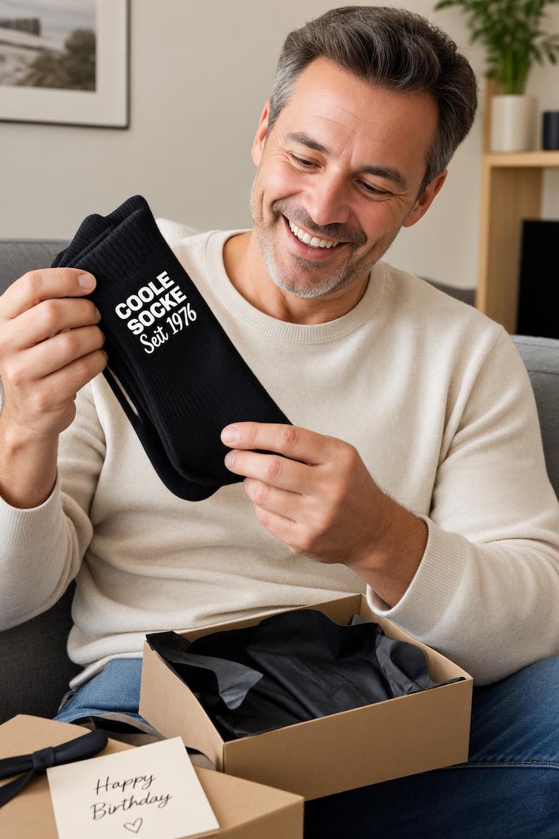 May include: A pair of black crew socks featuring the white printed text COOLE SOCKE Seit 1976. The socks are made of a ribbed fabric and are presented as a gift, accompanied by a small card with the handwritten text Happy Birthday and a heart symbol.