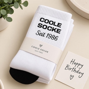 May include: A pair of white crew socks with a black heel and toe, featuring the black text COOLE SOCKE Seit 1986. The socks are wrapped in a beige paper band with the same text and a heart icon, accompanied by a card that says Happy Birthday.