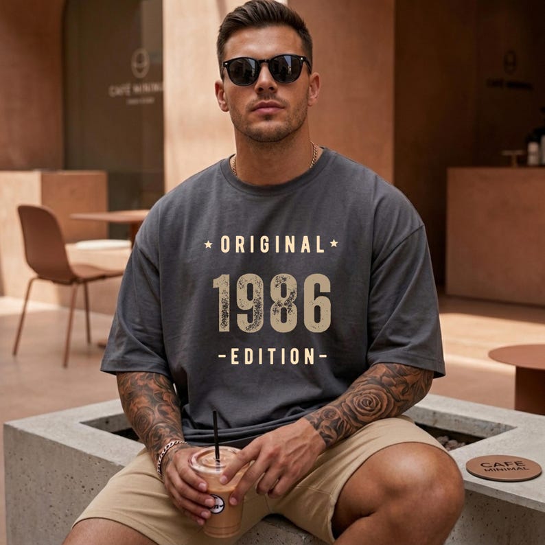 May include: A charcoal grey short-sleeved t-shirt featuring the text ORIGINAL 1986 EDITION in a light beige, distressed font. The shirt has a crew neckline and a relaxed fit, with the graphic centred on the chest between two small stars.