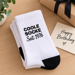 Könnte beinhalten: Ein Paar weiße Socken mit schwarzer Ferse und Spitze. Auf der Seite der Socken ist der Text "COOLE SOCKE Seit 1976" in Schwarz aufgedruckt. Die Socken sind gefaltet und liegen neben einer Karte mit der Aufschrift "Happy Birthday" und einem Herz.