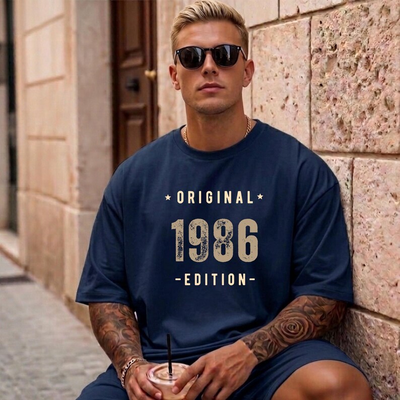 May include: A navy blue short-sleeved t-shirt featuring the text ORIGINAL 1986 EDITION in a light beige, distressed font. The design includes a small star on each side of the word ORIGINAL. The shirt has a crew neckline and a loose fit.