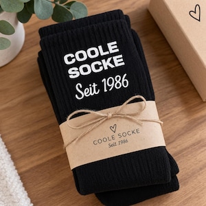 May include: A pair of black ribbed socks featuring the white printed text COOLE SOCKE Seit 1986. The socks are secured with a tan paper band that displays the same text and a small heart icon, tied together with a thin brown twine bow.