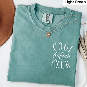 Puede incluir: Camiseta verde claro con la inscripción blanca "COOL AUNTS CLUB". La camiseta tiene cuello redondo y mangas cortas. Un collar plateado con un colgante redondo está sobre la camiseta.
