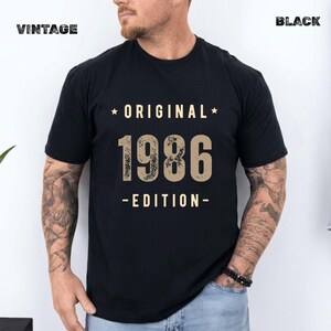 May include: A black short-sleeved t-shirt featuring the text "ORIGINAL 1986 EDITION" in a distressed beige font. The word "ORIGINAL" is centred above the large numbers "1986," with "EDITION" below, all flanked by small stars.