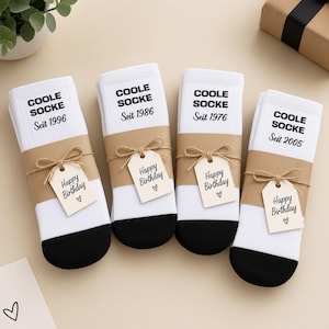 May include: Four pairs of white crew socks with black toes, each wrapped in a brown paper band. The socks feature the black text COOLE SOCKE and a birth year, with a small brown tag tied on that reads Happy Birthday with a heart symbol.