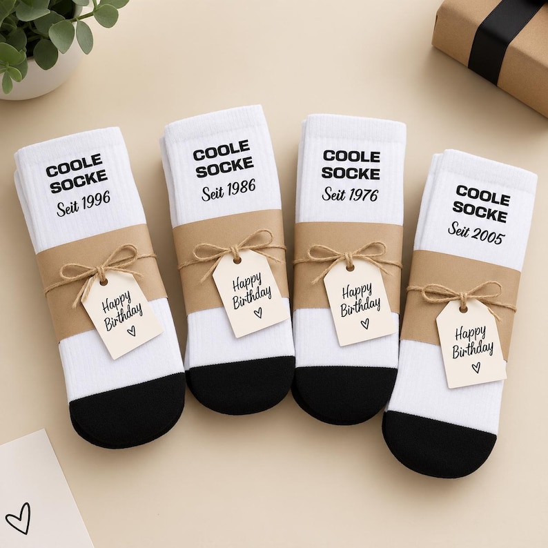 May include: Four pairs of white crew socks with black toes, each wrapped in a brown paper band. The socks feature the black text COOLE SOCKE and a birth year, with a small brown tag tied on that reads Happy Birthday with a heart symbol.