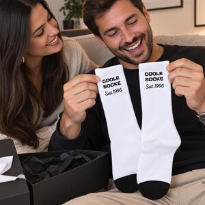 May include: A pair of white crew socks with black heels and toes. The text COOLE SOCKE is printed in a bold black sans-serif font, followed by the text Seit 1996 in a black serif font on the upper portion of each sock.