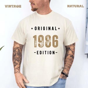 May include: A cream-coloured short-sleeve t-shirt featuring the black text ORIGINAL and EDITION with a star on each side of the word ORIGINAL. The centre of the shirt displays the large, distressed-style tan numbers 1986.