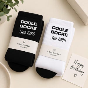 May include: A pair of ribbed socks, one black and one white, each featuring the text COOLE SOCKE Seit 1986. Both socks are wrapped in a paper band with the same text and a heart icon. A small card nearby displays the text Happy Birthday and a heart.