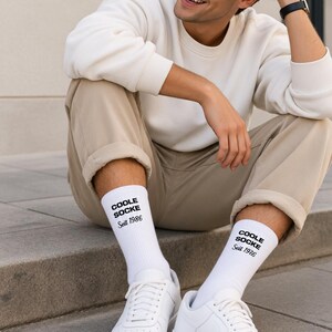 May include: A pair of white crew socks featuring the black text COOLE SOCKE Seit 1986 printed on the outer side of each sock. The socks are made of a ribbed knit fabric and are worn with white sneakers, a cream sweatshirt, and beige trousers.