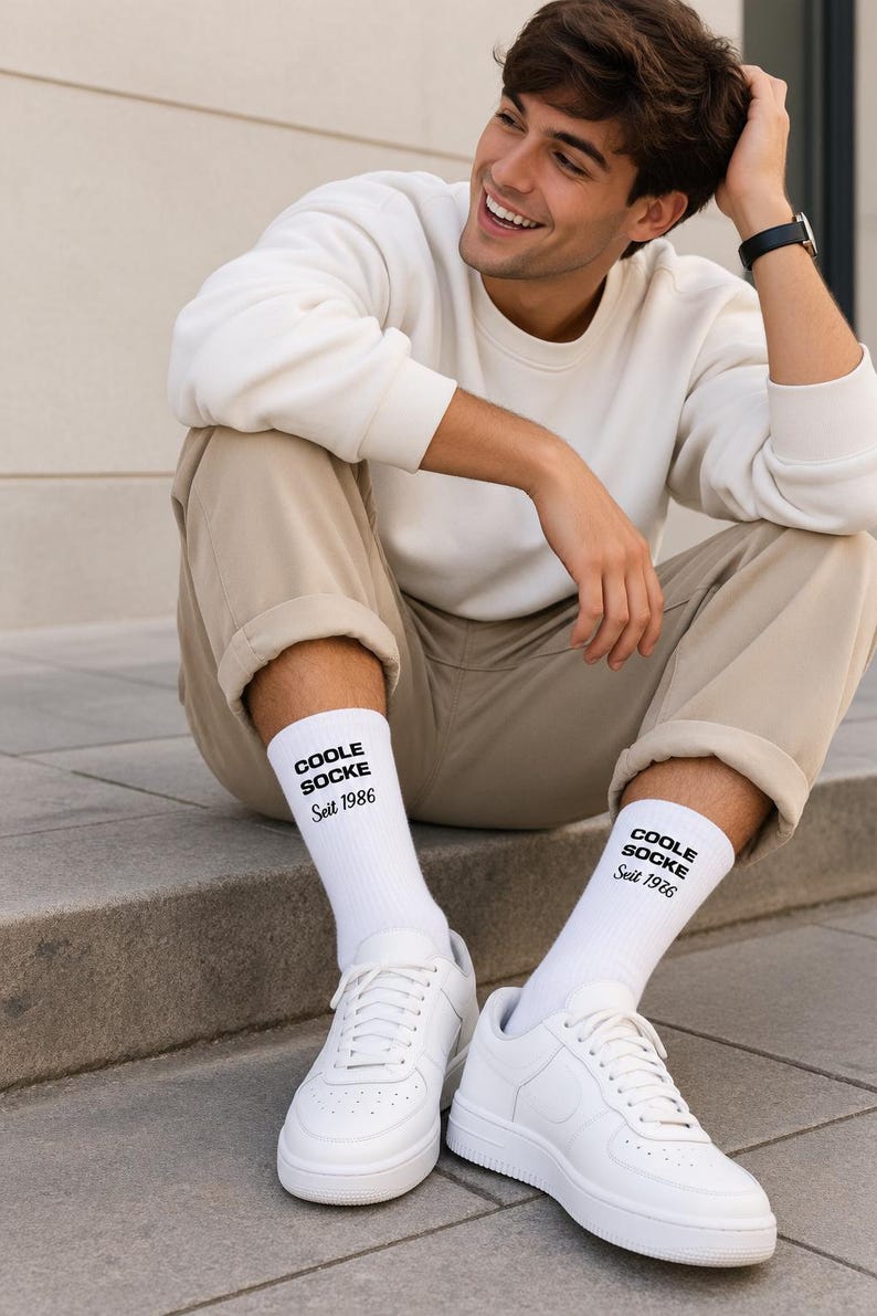 May include: A pair of white crew socks featuring the black text COOLE SOCKE Seit 1986 printed on the outer side of each sock. The socks are made of a ribbed knit fabric and are worn with white sneakers, a cream sweatshirt, and beige trousers.