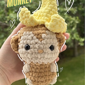 May include: A handmade crochet monkey with a banana hat. The monkey is primarily beige and brown, with black safety eyes. The text "Banana Monkey" is in green and yellow. The image also includes the text "PDF Crochet Pattern".