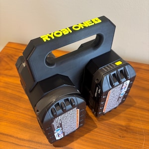 May include: Black and yellow Ryobi One+ battery holder with two black batteries. The holder has a handle and the Ryobi One+ logo in yellow. The batteries have warning labels.