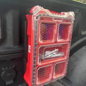Peut inclure: Une boîte de rangement rouge Milwaukee Packout avec des compartiments transparents en forme de nid d'abeille. La boîte est montée sur une surface noire, avec le nom de la marque visible. Le couvercle est transparent.