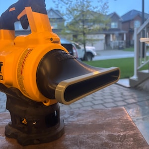 May include: A yellow and black leaf blower with a black nozzle. The blower is sitting on a brown surface. The handle is black with an orange accent. The background shows a residential neighborhood.