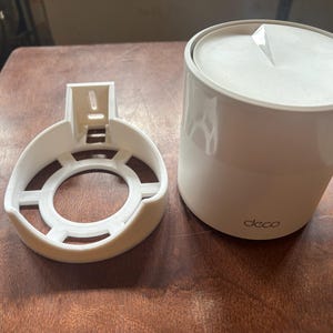 May include: A white wall mount for a Deco router. The mount is made of plastic and has a circular opening in the center. The router is not included.