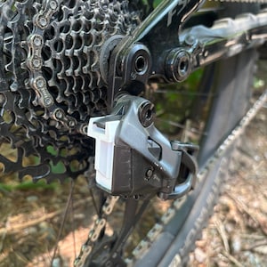 May include: Close-up of a black bicycle derailleur with a white plastic cage. The derailleur is attached to a black bicycle frame with a silver chain.
