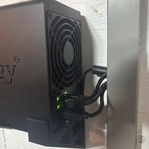 May include: A black box with a fan and a green ethernet cable plugged into it. The box is mounted to a white wall.