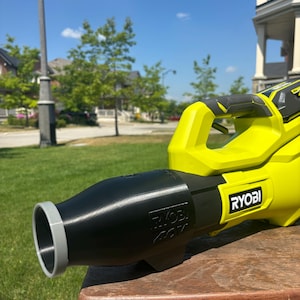May include: A bright yellow and black Ryobi leaf blower sits on a wooden surface. The blower has a black nozzle with a gray rim and the Ryobi logo is visible. The background shows a green lawn, trees, and a blue sky.