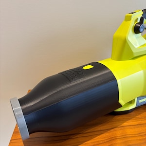 May include: A cordless leaf blower, featuring a black nozzle and a bright yellow body. The blower is resting on a wooden surface. The nozzle has the brand name printed on it. The blower is designed for outdoor use.