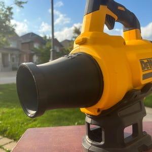 May include: A yellow and black cordless leaf blower with a black nozzle and a black base. The blower is on a brown wooden surface.