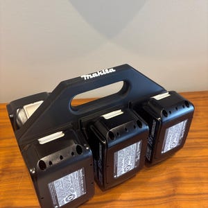 May include: A black plastic battery holder with a handle that says "Makita" on it. The holder has four slots for batteries and is holding three black batteries.