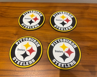 May include: Four circular coasters featuring the Pittsburgh Steelers logo. Each coaster displays the team's logo: a yellow star, a red star, and a black star, with the words "Pittsburgh Steelers" in a circular design.