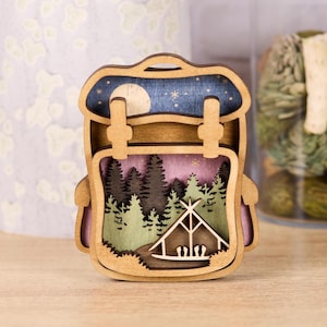 May include: A wooden backpack-shaped decoration with a layered design. The top layer depicts a night sky with a moon and stars. The middle layer shows a forest scene with a tent. The color palette includes shades of brown, blue, pink, and green.