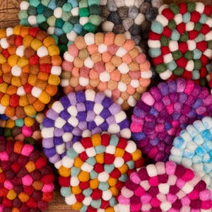 May include: A collection of colorful, round felt coasters in various shades of pink, purple, orange, red, green, and blue. Each coaster is made of small, tightly packed felt balls, creating a textured surface. The coasters are arranged on a wooden surface.