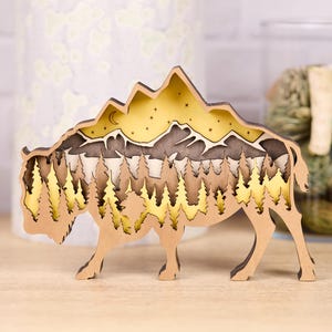May include: A wooden bison-shaped art piece with a layered design depicting a mountain landscape with trees, a moon, and stars. The bison is light brown, and the landscape has shades of brown and yellow. The art piece is on a wooden surface.