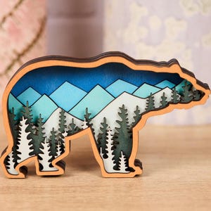 May include: A wooden bear-shaped decorative item with a layered design. The interior features a mountain and forest scene in shades of blue, green, and white. The bear's outline is a warm orange color, creating a striking contrast.