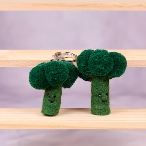 Puede incluir: Dos llaveros de fieltro verde en forma de brócoli con caras sonrientes. Los llaveros están colgados de un estante de madera.
