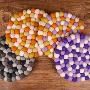 Op de afbeelding: Een verzameling ronde vilten onderzetters in verschillende kleuren, waaronder grijs, oranje, geel, roze, paars en wit. Elke onderzetter is gemaakt van kleine, dicht opeengepakte vilten ballen. De onderzetters liggen op een houten oppervlak.