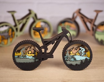 May include: A layered wooden mountain bike sculpture featuring a dark frame and wheels with intricate cutouts of cyclists and mountain landscapes inside the tires. Two additional similar sculptures in light green and brown are visible behind.