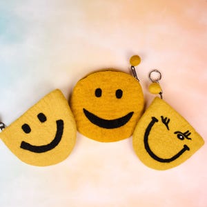 May include: Three yellow felt pouches with black embroidered smiley faces. The pouches are shaped like half circles and have a small black pom-pom on the top.