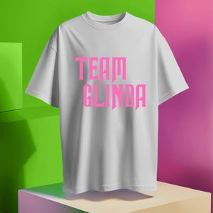 May include: White t-shirt with pink text that reads "Team Glinda".