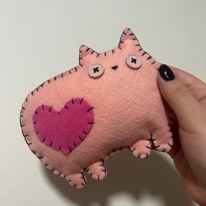 May include: A pink felt cat-shaped plush toy with a stitched black outline. The cat has button eyes and a pink heart applique. The toy is handmade and has a whimsical, cute design.