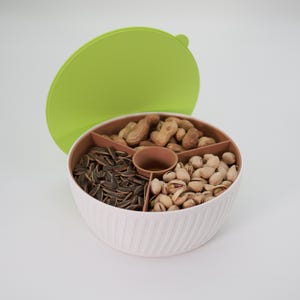 Puede incluir: Un bol de aperitivos blanco y verde de cuatro secciones con tapa. El bol está lleno de pistachos, cacahuetes y semillas de girasol.