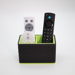 Può includere: Un organizer per telecomandi nero e verde che contiene tre telecomandi. L'organizer ha un design a coste sull'esterno nero e un interno verde brillante. I telecomandi sono bianchi e neri.