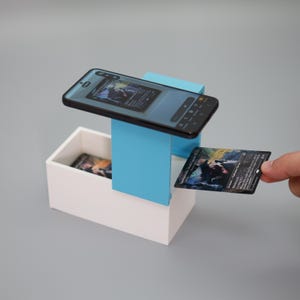K&ouml;nnte beinhalten: Ein Smartphone ist &uuml;ber einer wei&szlig;en Box und einer blauen Halterung positioniert und zeigt ein Kartenbild. Eine Person h&auml;lt eine Sammelkarte, mit einer &auml;hnlichen Karte in der Box. Die Karten haben einen dunklen Hintergrund mit farbenfrohen Illustrationen.