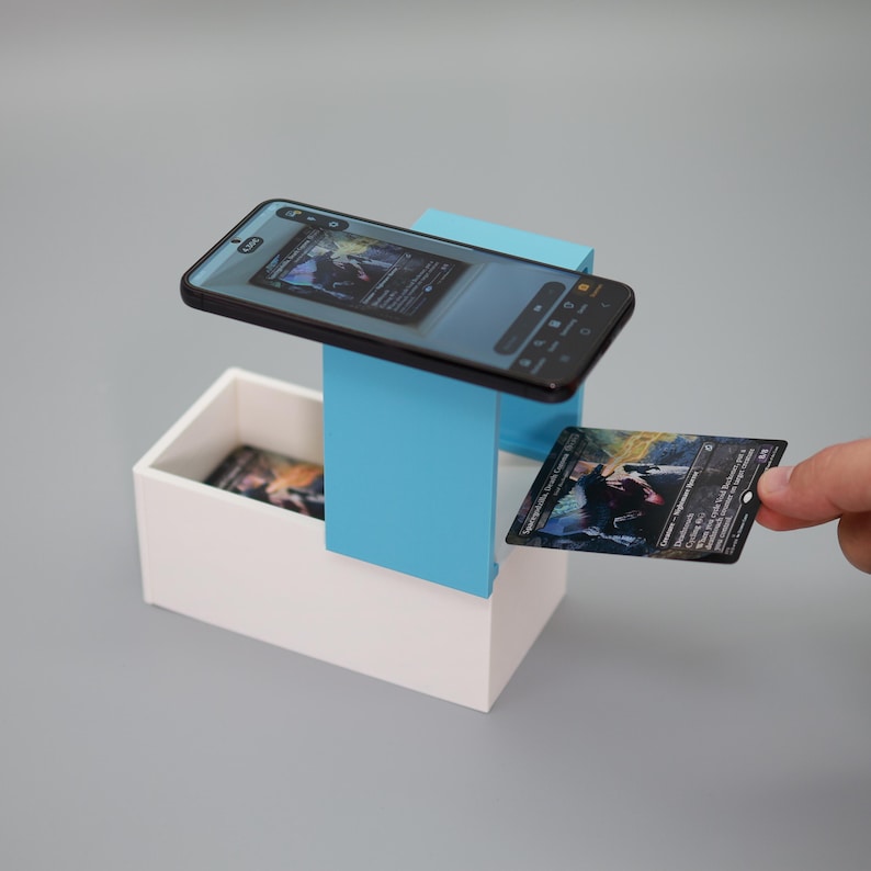 K&ouml;nnte beinhalten: Ein Smartphone ist &uuml;ber einer wei&szlig;en Box und einer blauen Halterung positioniert und zeigt ein Kartenbild. Eine Person h&auml;lt eine Sammelkarte, mit einer &auml;hnlichen Karte in der Box. Die Karten haben einen dunklen Hintergrund mit farbenfrohen Illustrationen.