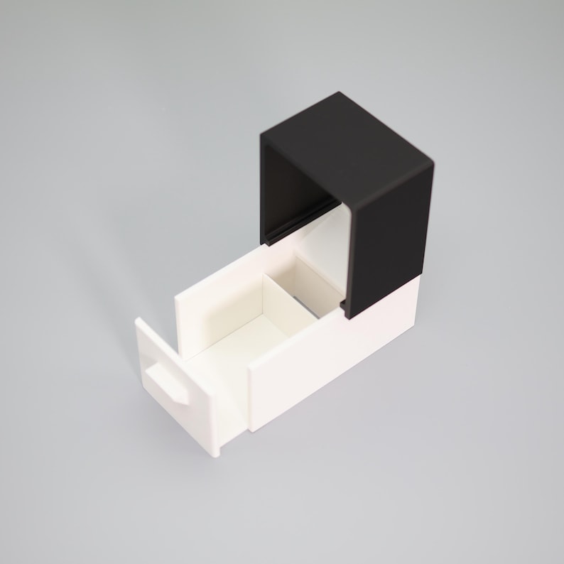 K&ouml;nnte beinhalten: Eine moderne, zweifarbige Aufbewahrungsbox mit schwarzem Deckel und wei&szlig;em Sockel. Die Box hat eine Schublade, die teilweise ge&ouml;ffnet ist und ein unterteiltes Innere freilegt. Das Design ist minimalistisch und geometrisch.