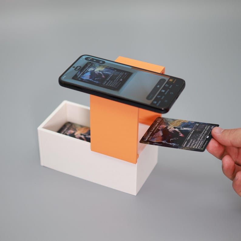 K&ouml;nnte beinhalten: Ein wei&szlig;-oranger Kartenst&auml;nder mit Smartphone und Sammelkarten. Das Smartphone ist &uuml;ber dem St&auml;nder abgewinkelt und zeigt ein Kartenbild an. Eine Hand h&auml;lt eine Karte, weitere Karten befinden sich in der wei&szlig;en Box.